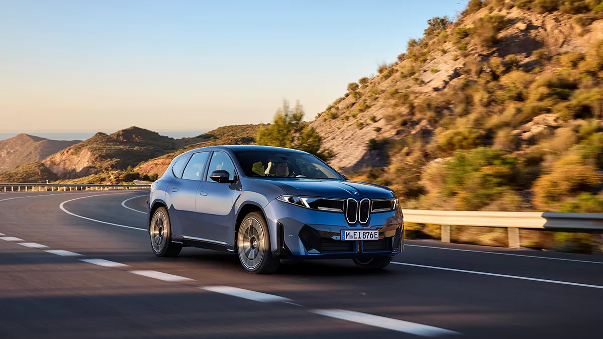 Blue BMW SUV driving along a winding coastal highway with rocky hills in the background at sunset light.