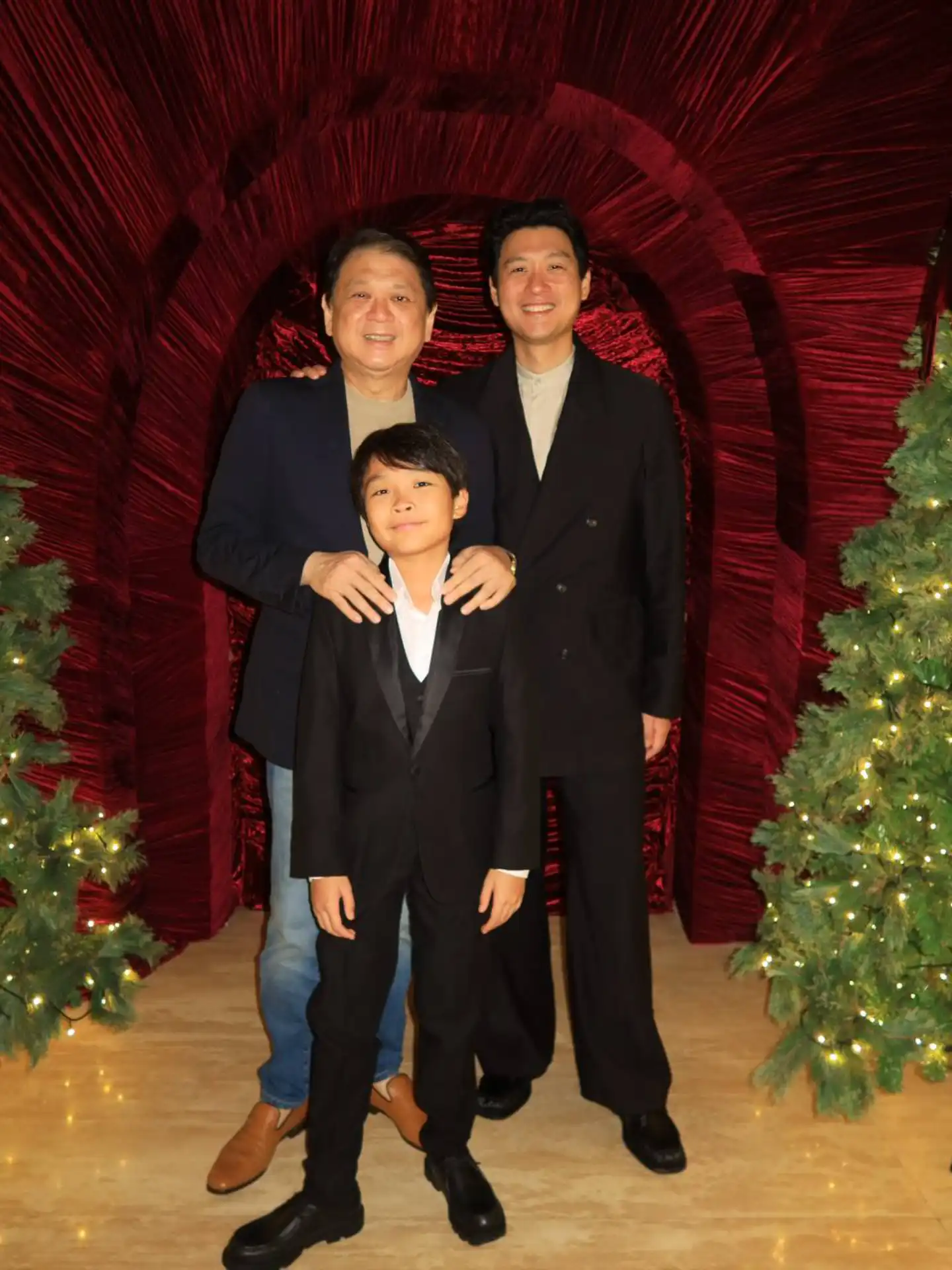 Three people in formal outfits posing for a photo in front of a red velvet arch and Christmas trees with lights at a holiday display.