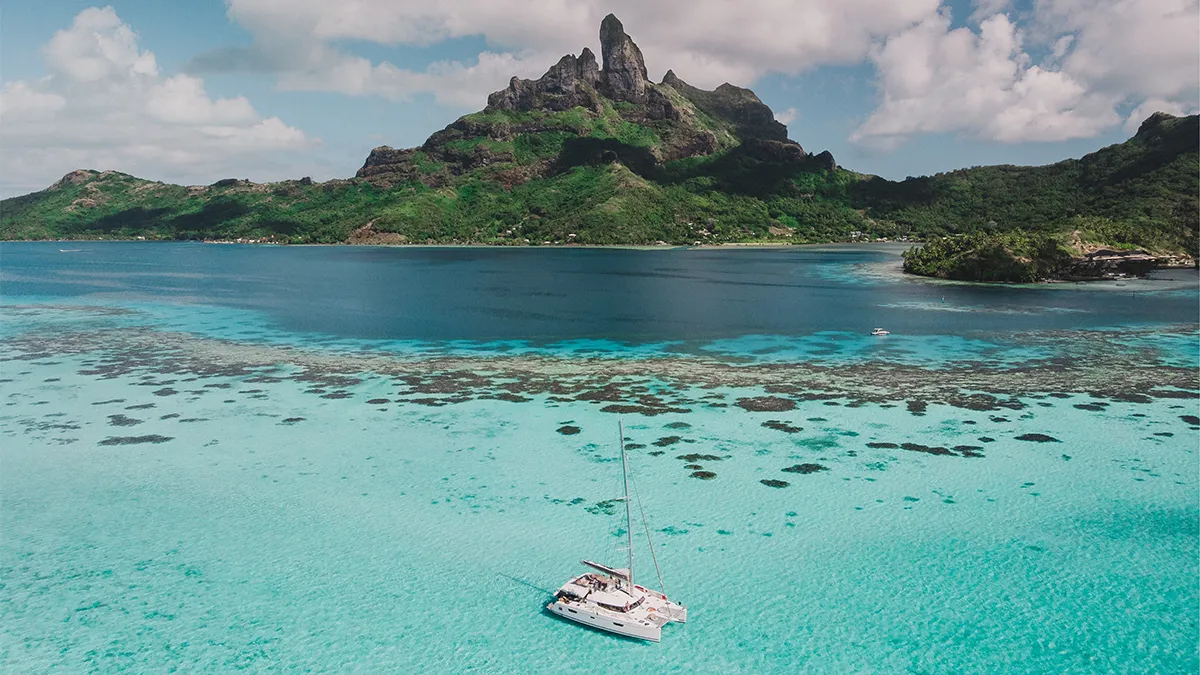 Sailboat anchored in bright turquoise lagoon with a lush green island and jagged mountain in the background under a partly cloudy sky