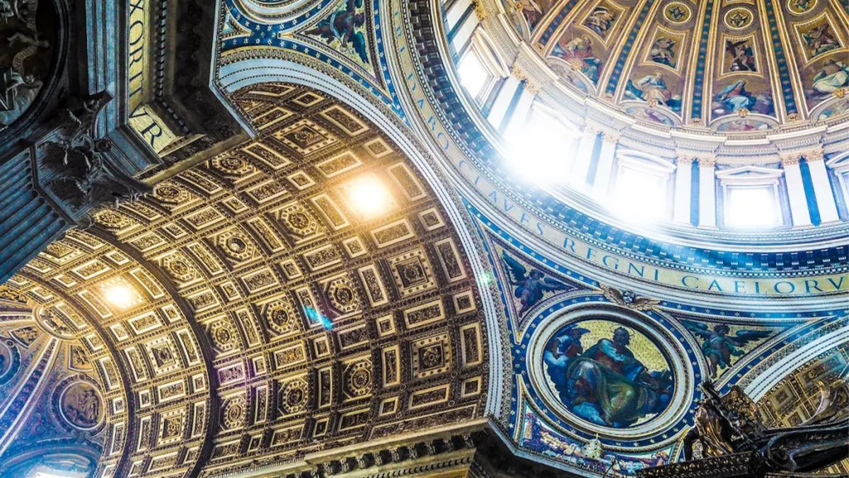 Interior of a grand cathedral dome with gold and blue mosaics, intricate coffering, and arched windows letting in light.