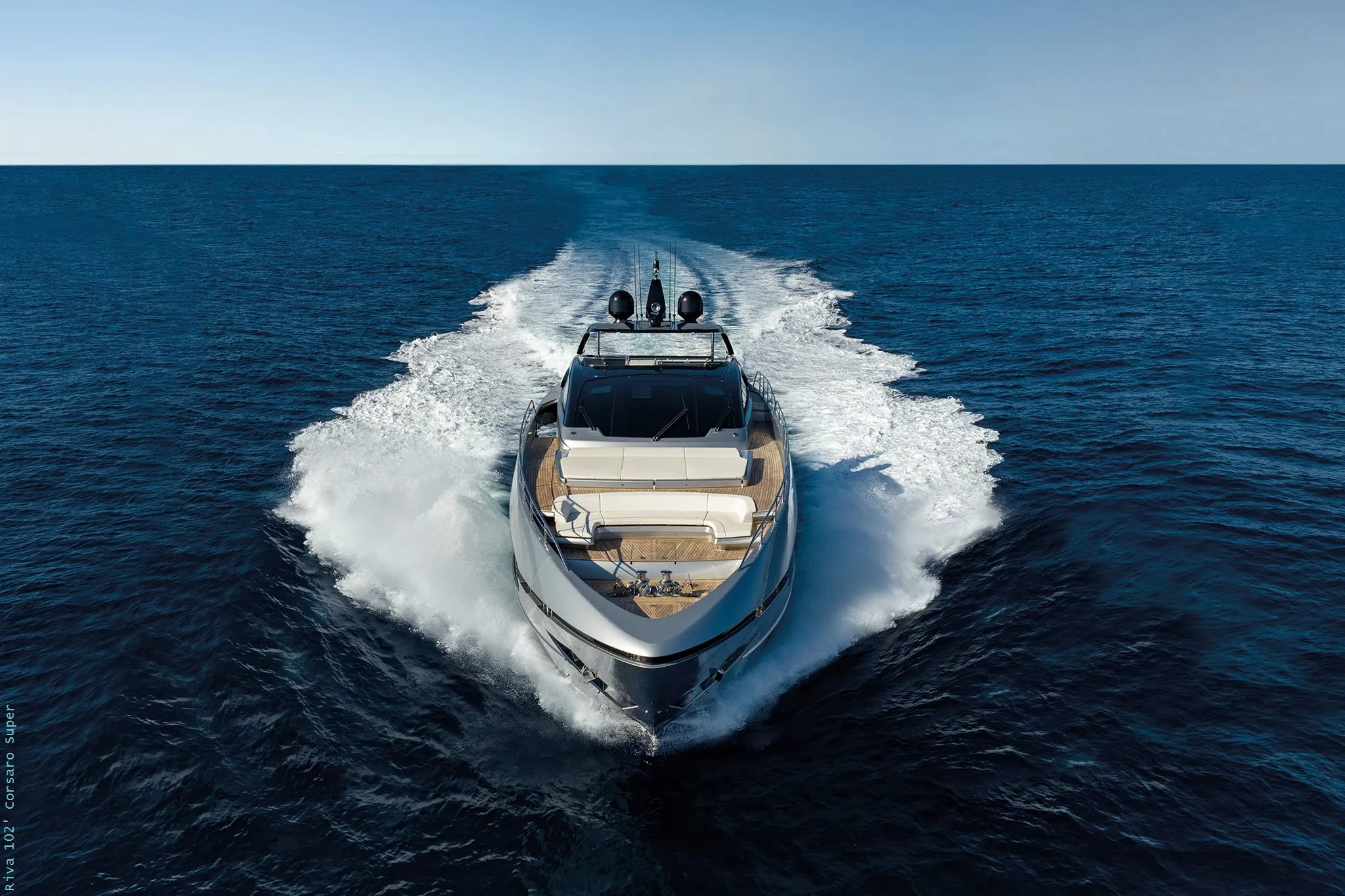 A sleek luxury motor yacht viewed from above, traveling forward through deep blue water with white wake trailing behind.
