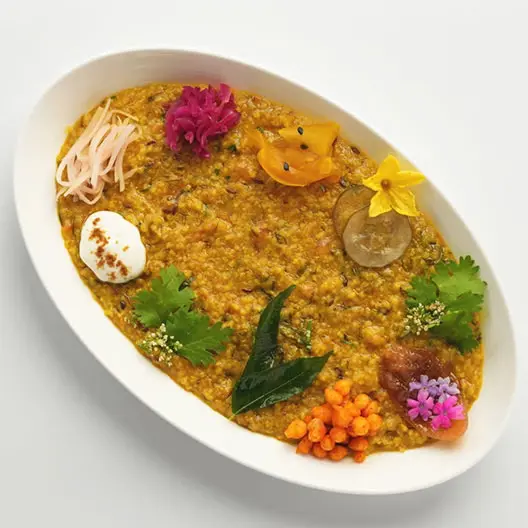 Oval white bowl containing yellow-orange curry, garnished with cilantro, edible flowers, a halved boiled egg, lime slice, and assorted colorful garnishes.