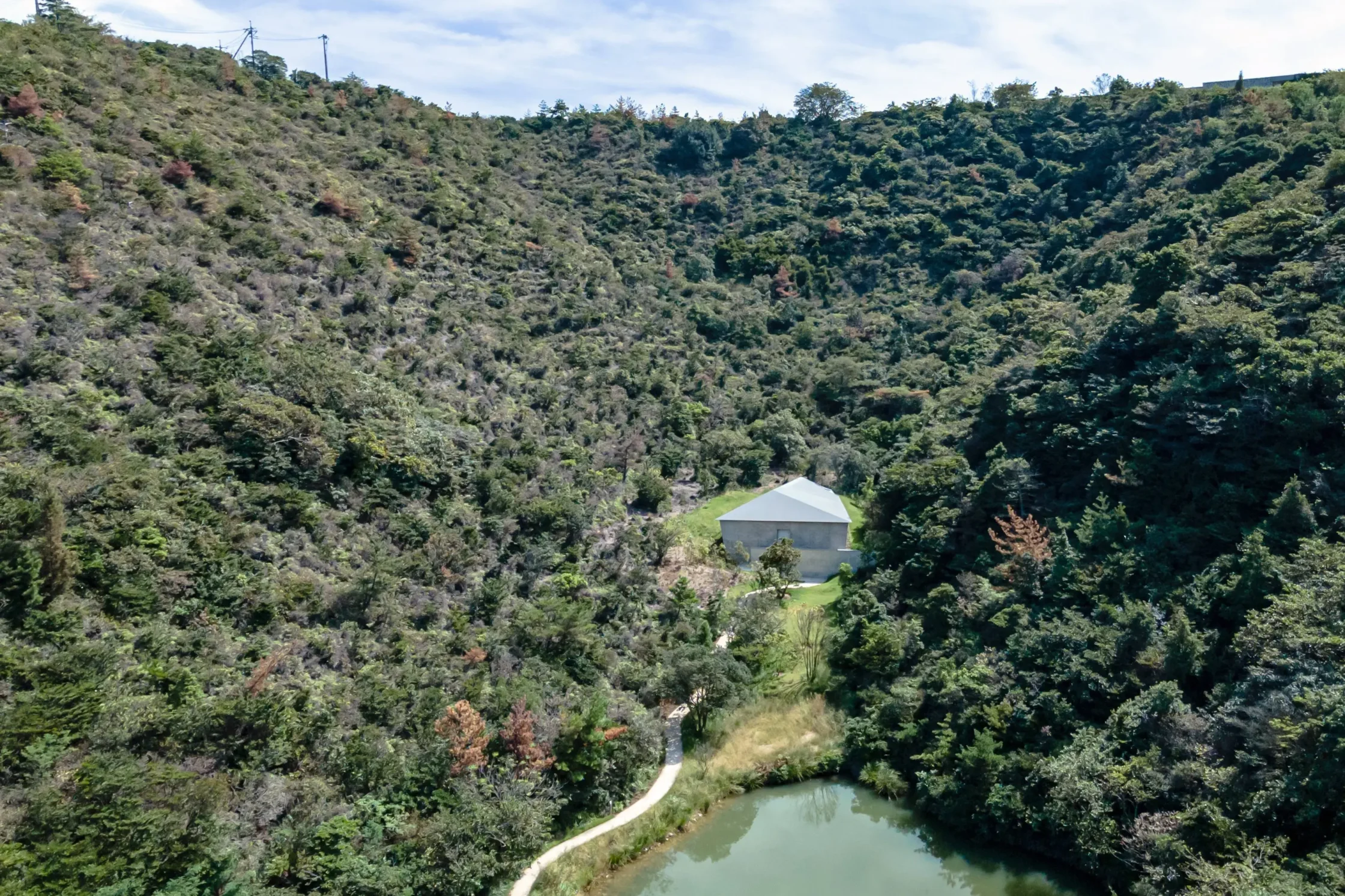 Dense forested hillside with a small gray building and white roof, a winding path, and a river at the bottom.
